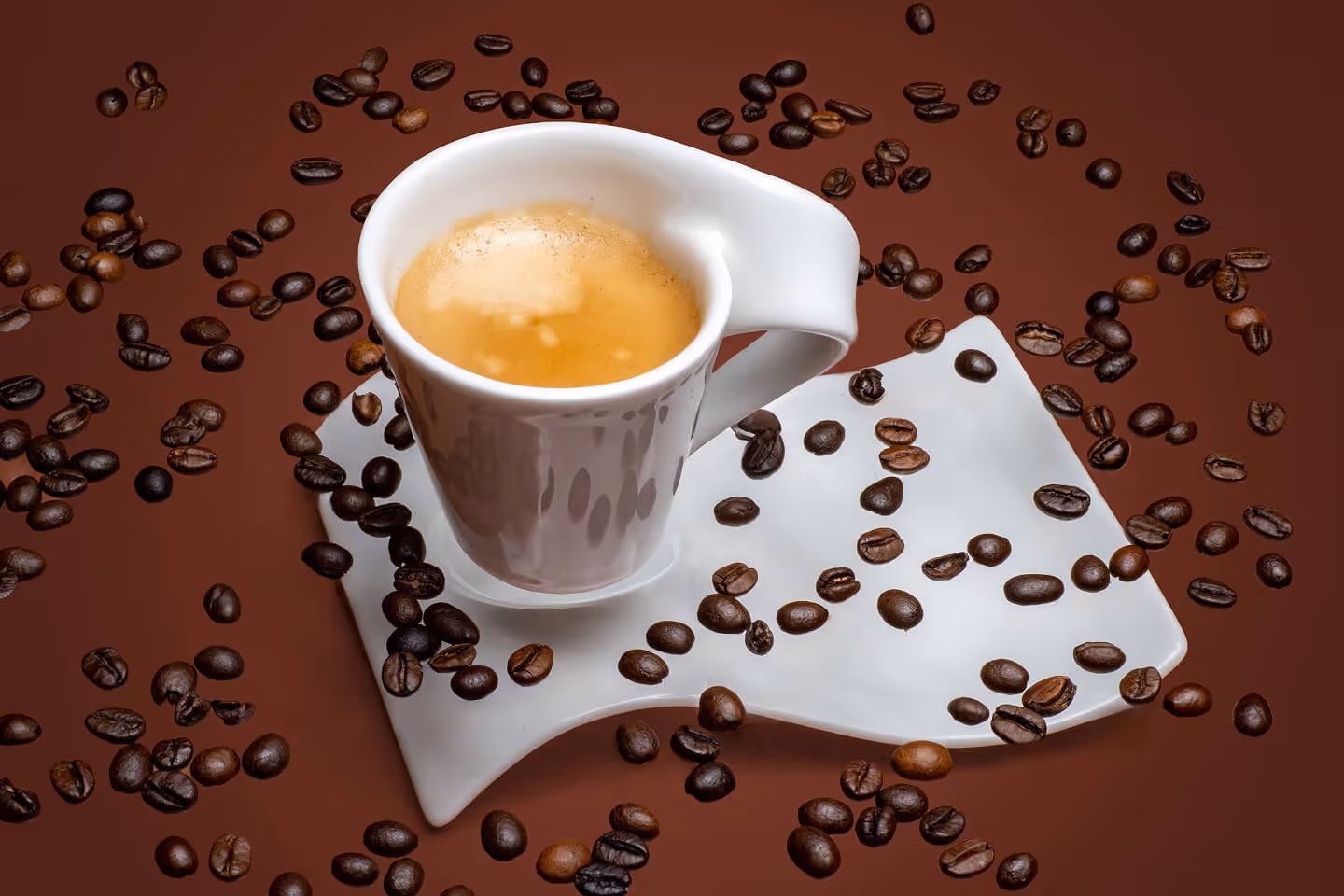 Espresso in a unique white cup and saucer, topped with rich crema and surrounded by roasted coffee beans on a brown surface.