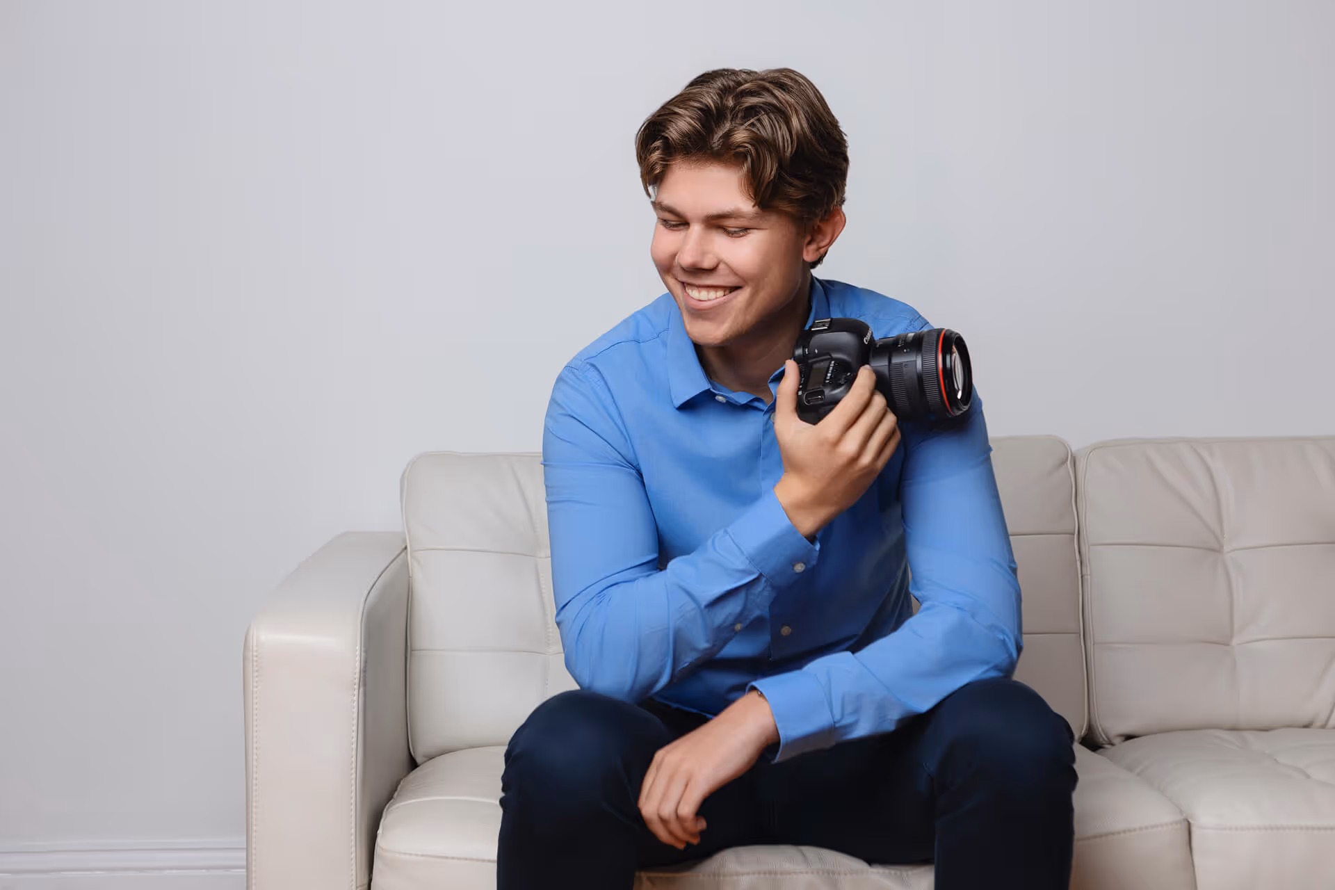 Lars in a blue shirt sitting on a white sofa, smiling while holding a DSLR camera. The atmosphere is relaxed and joyful.
