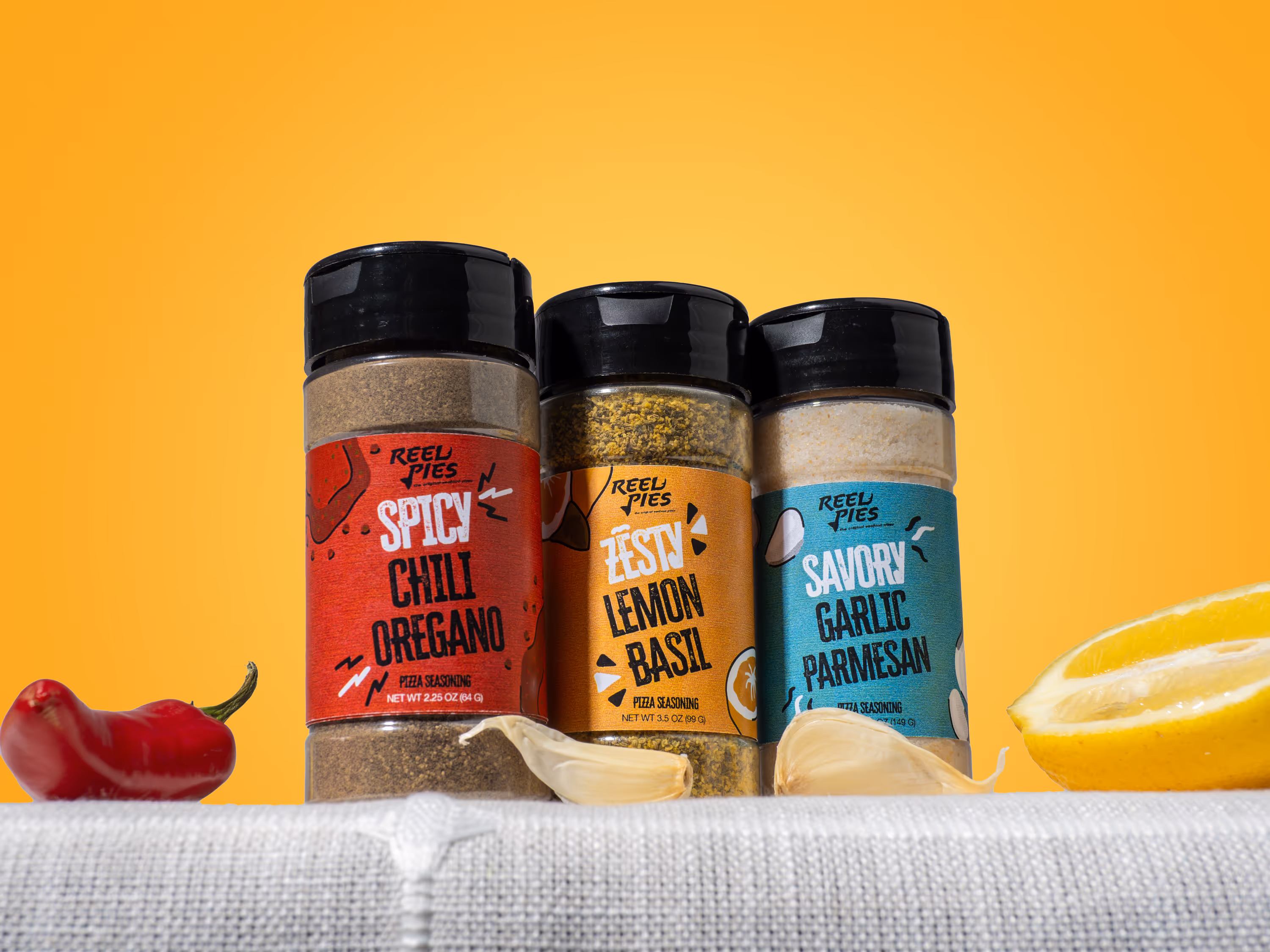 Reel Pies pizza seasoning variety pack featuring Spicy Chili Oregano, Zesty Lemon Basil, and Savory Garlic Parmesan flavors in shaker jars with fresh ingredients.