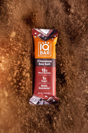 A chocolate sea salt protein bar with a rich brown and orange wrapper featuring text highlighting 12g plant protein and brain nutrients, set on a cocoa powder background.