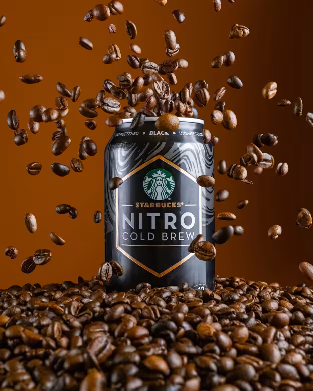 A can of Starbucks Nitro Cold Brew Black, Unsweetened, with coffee beans exploding around and below it against a brown background.