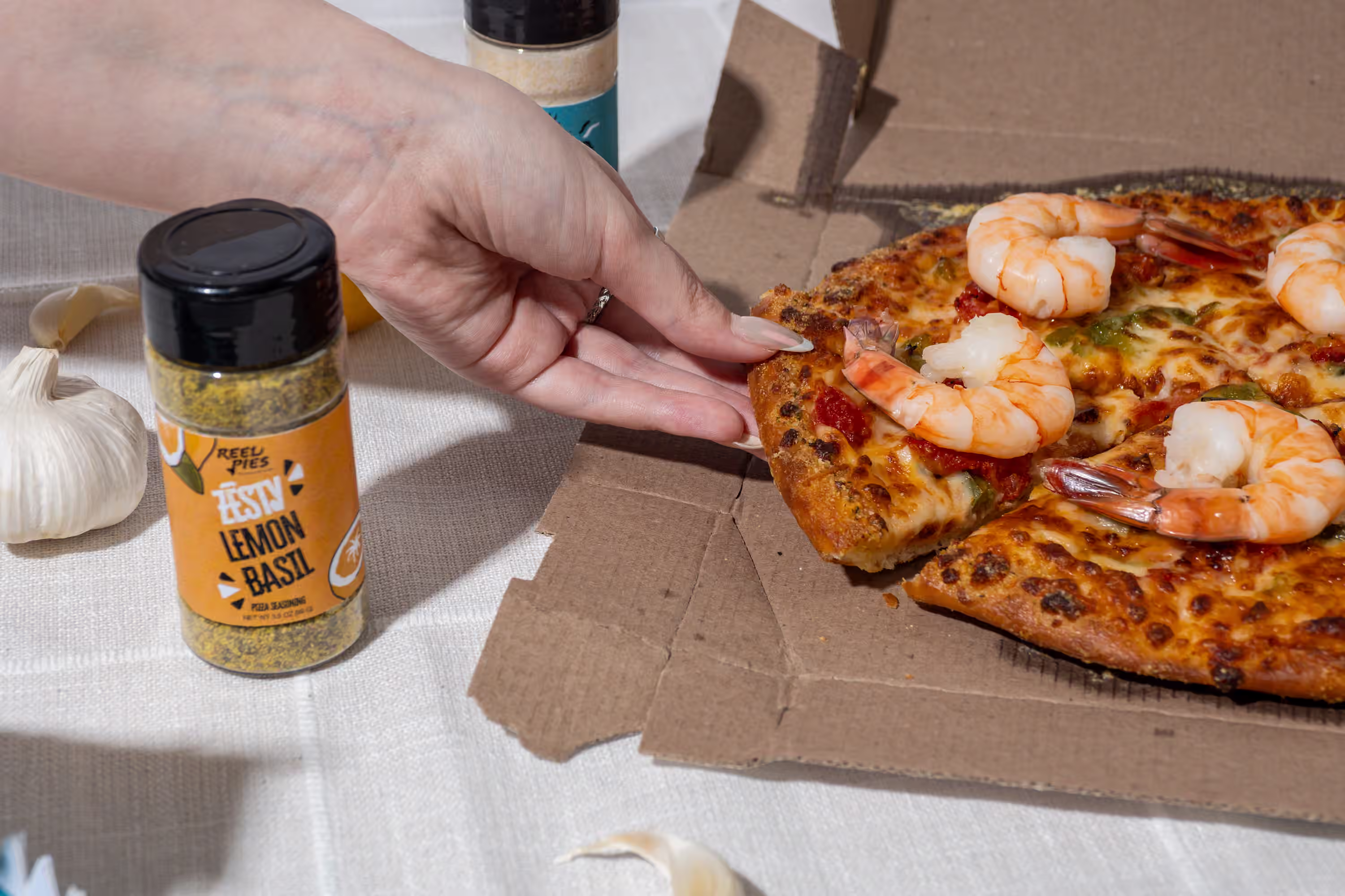 Reel Pies Zesty Lemon Basil pizza seasoning placed next to a shrimp pizza with a hand grabbing a slice in a lifestyle product photograph