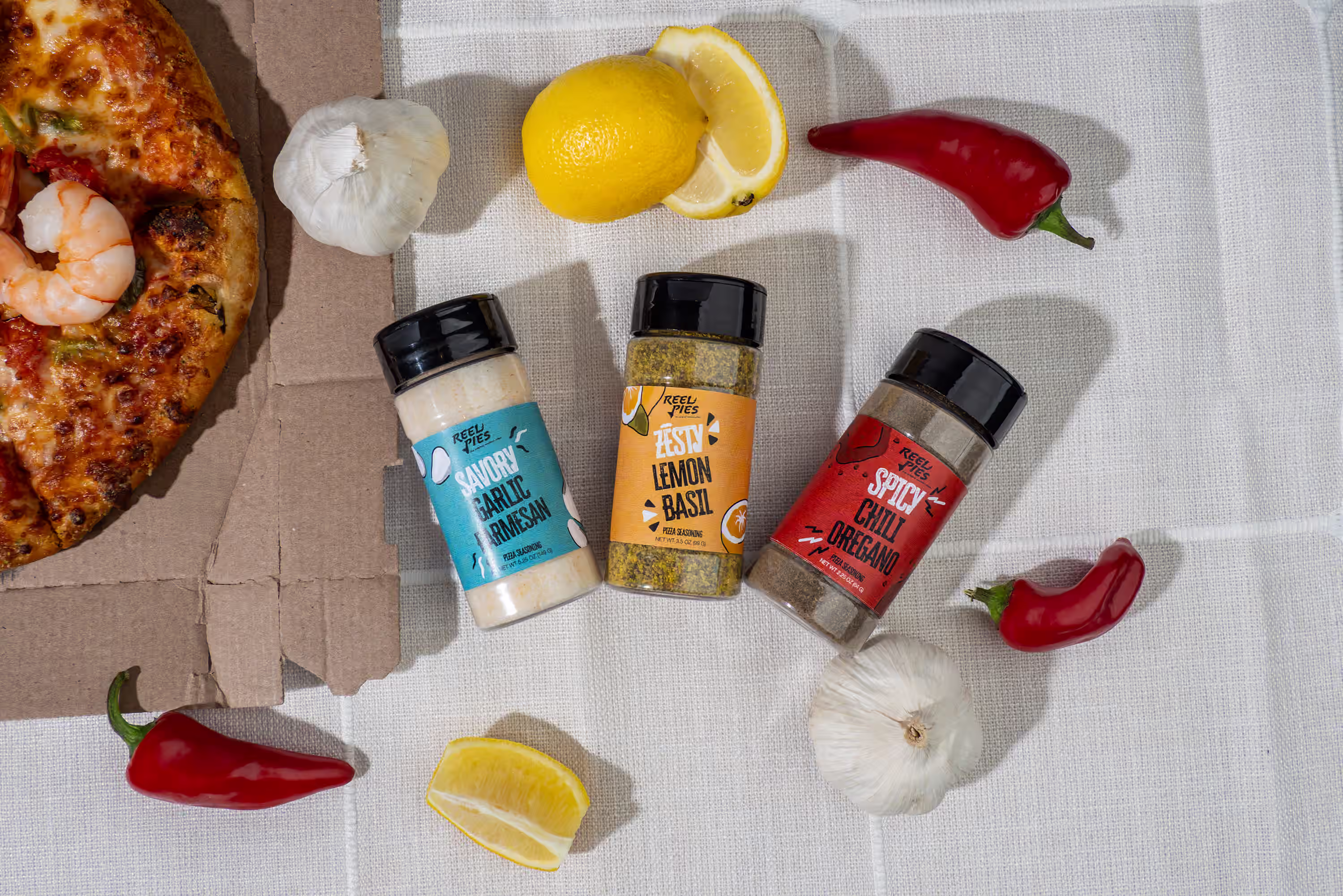 Overhead flat lay of three Reel Pies pizza seasoning flavors styled with fresh peppers, garlic, lemons, and pizza on a white linen surface