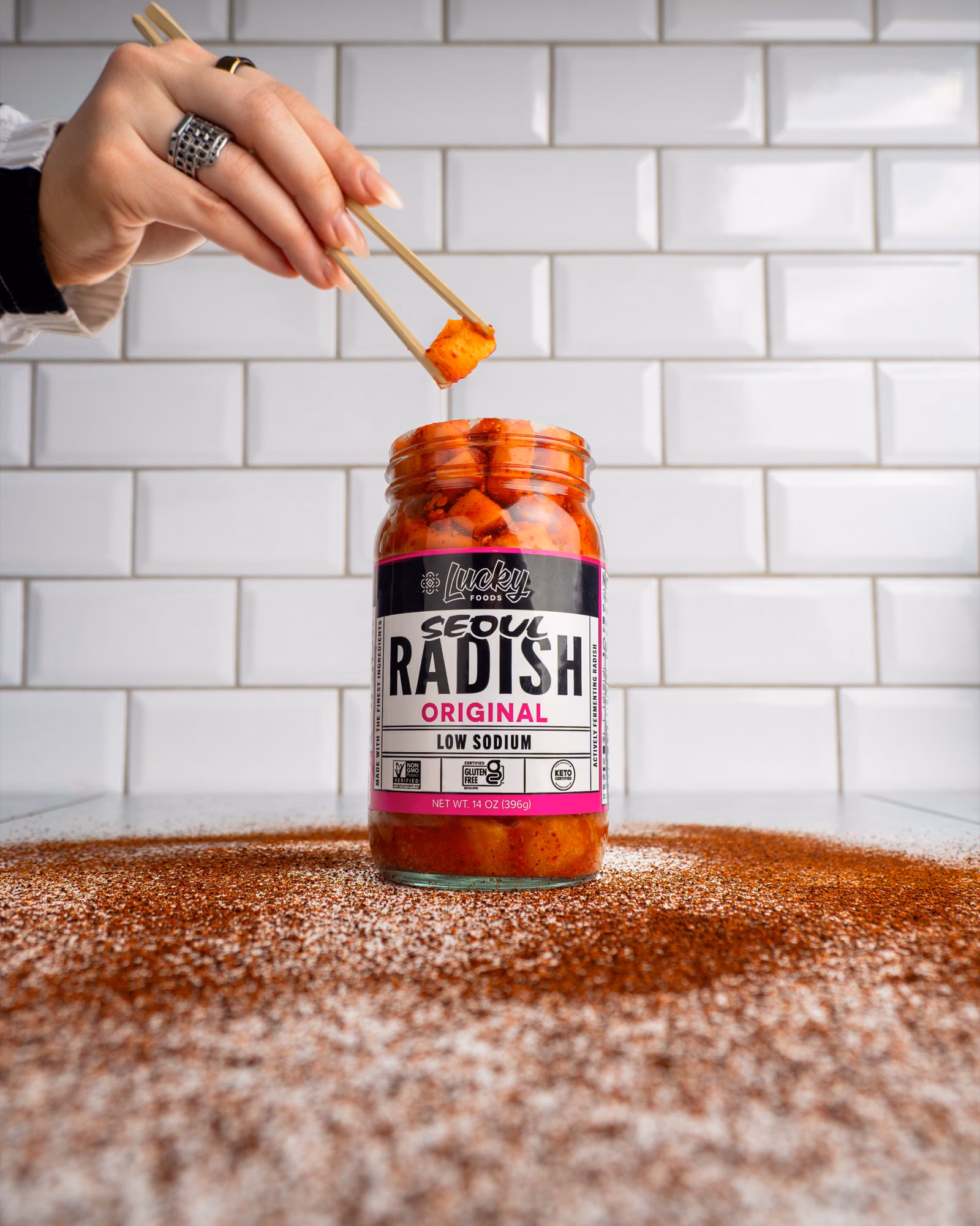 Chopsticks lifting a piece of Lucky Foods Seoul Radish Kimchi from an open jar in front of a white tile background.
